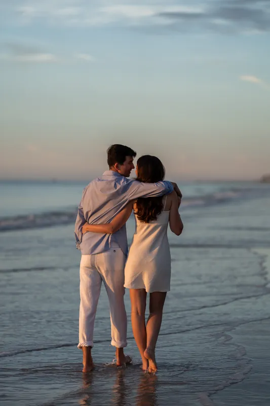 Sally and Pasha walk arm-in-arm through shallow beach water in matching cream linen clothing at dusk.