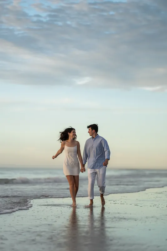 Sally and Pasha stroll hand-in-hand along the wet sand with calm ocean and cloudy sky at golden hour.