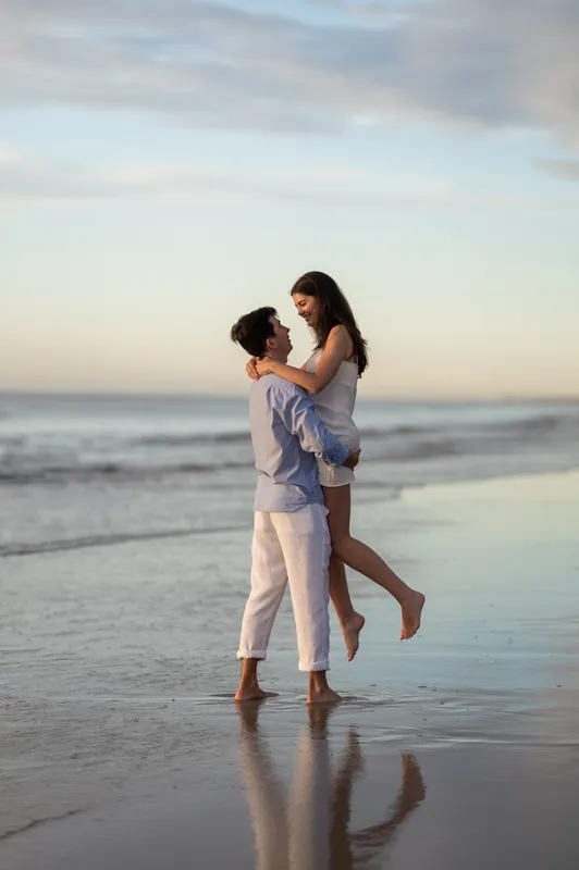 Pasha lifts Sally off the ground while standing in shallow water on the beach at sunset.