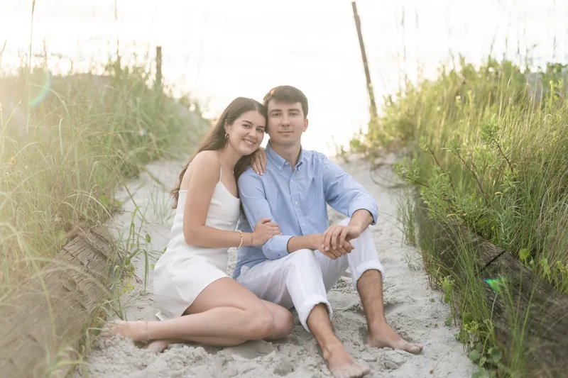 Sally and Pasha sit together on sandy beach path surrounded by beach grass at golden hour.