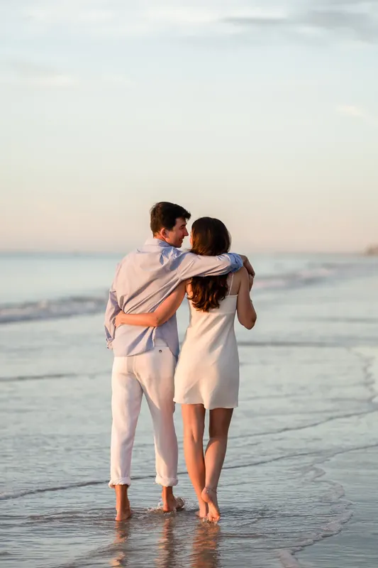 Sally and Pasha walk hand-in-hand through shallow waves at St. Augustine Beach in cream-colored linen, embracing from behind.