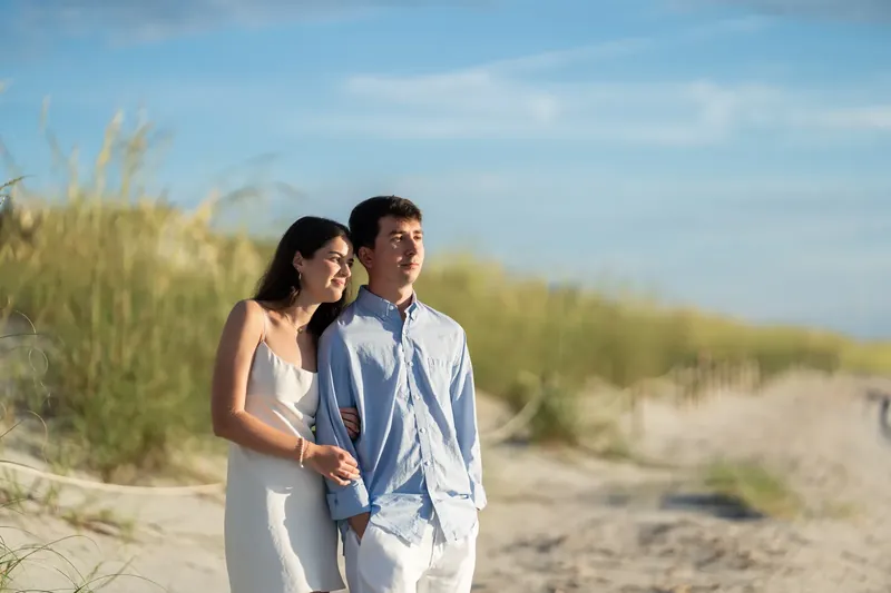 The couple strolls side by side through beach grass under a bright blue sky.