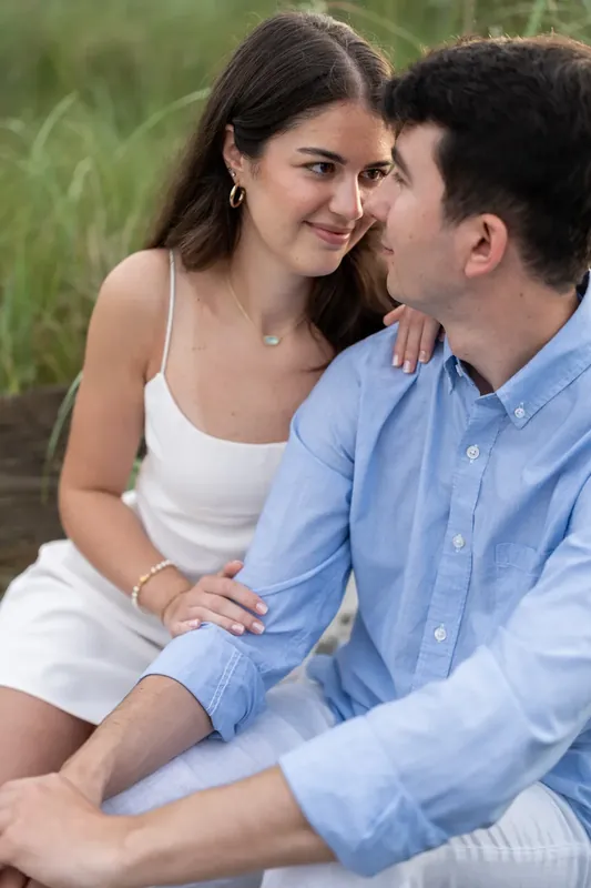 Sally smiles warmly while Pasha looks at her, both seated on the beach with dune vegetation behind them.