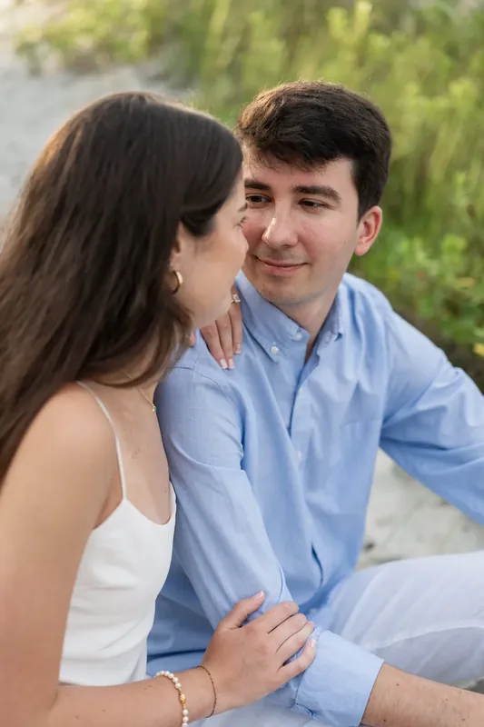 Pasha grins as Sally gazes at him, their hands clasped while sitting on the sand.