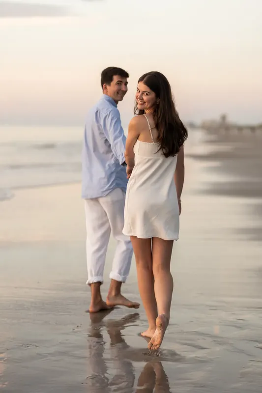 Both turned toward the ocean, Sally links her arm through Pasha's as dune grasses frame the beach behind them.