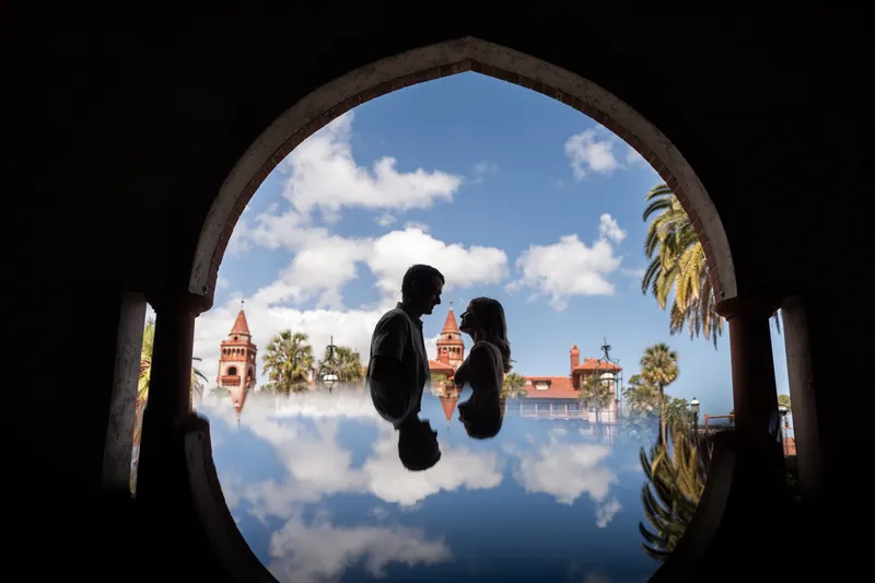 Samantha and Dillon kiss under a stone archway framing a reflection of downtown St. Augustine's historic buildings and blue sky.