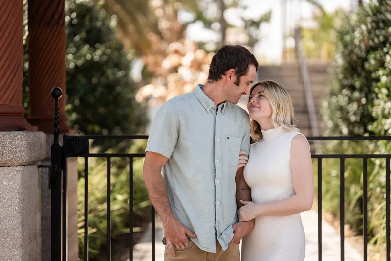 Dillon in a light blue button-up shirt and Samantha in a white sleeveless dress stand together on a balcony with palm trees and historic architecture visible behind them.