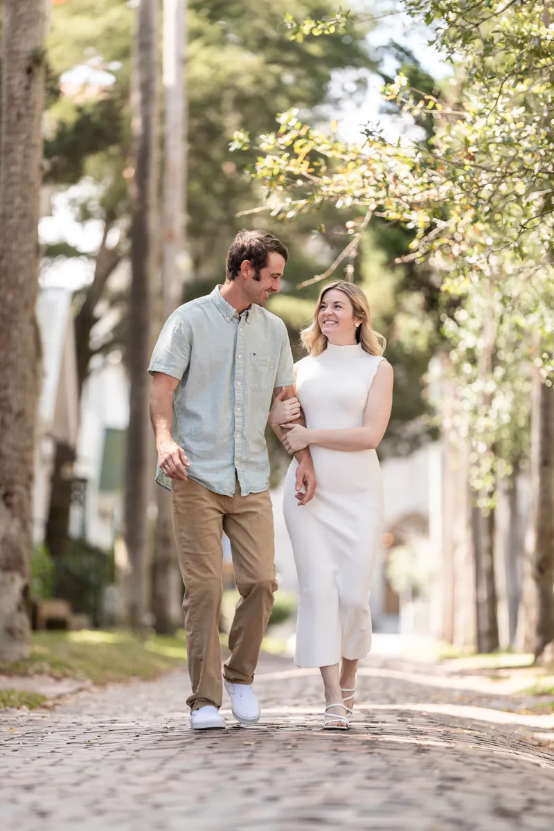 Samantha and Dillon walk together down a tree-lined brick pathway in downtown St. Augustine, holding hands.