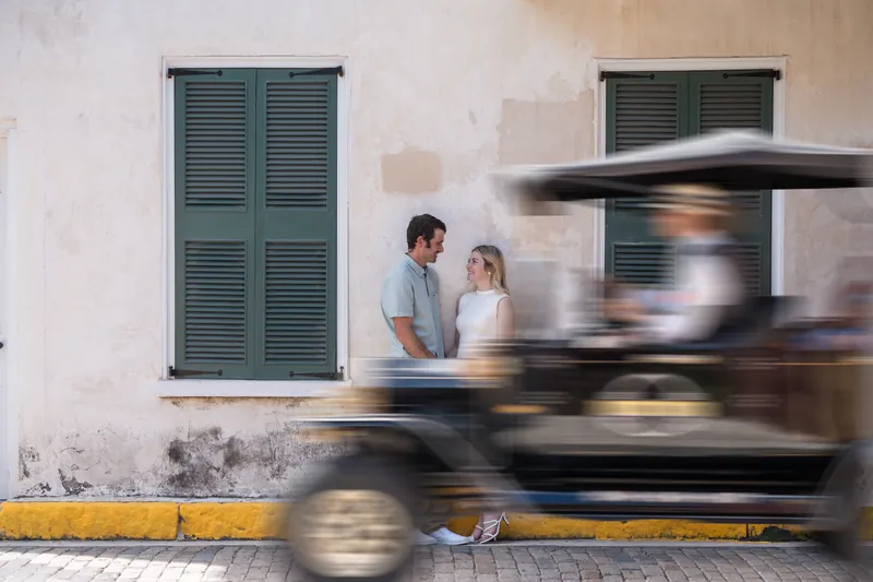 Samantha and Dillon stand against a weathered white building with green shutters as a horse-drawn carriage passes by on the street.