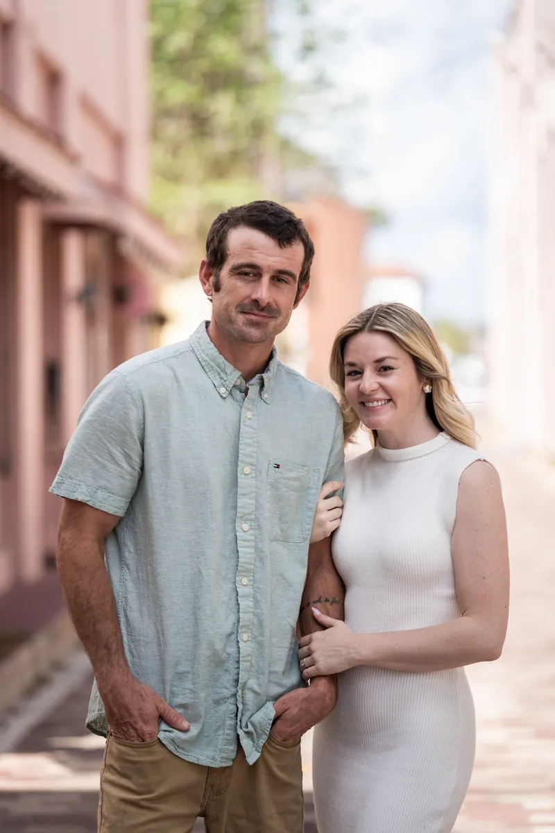 Dillon in a light blue button-up shirt and Samantha in a white sleeveless dress smile on a brick-lined downtown street.