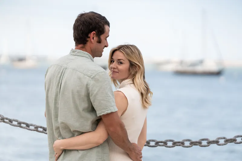 Samantha looks back at Dillon as they embrace against a chain railing with calm water and boats behind them.