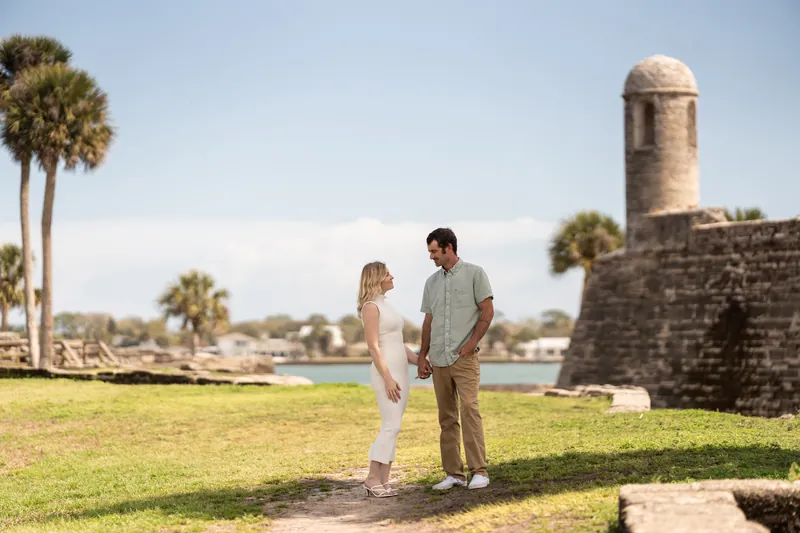 Dillon and Samantha stand on the grass near the historic coquina fort with palm trees and water visible in the background.
