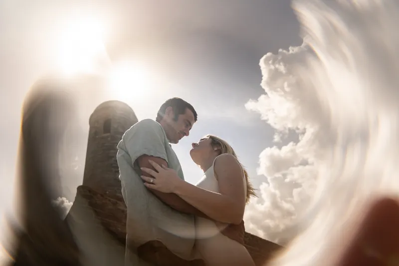 Samantha and Dillon embrace outdoors with bright sunlight and dramatic clouds creating a backlit effect around them.