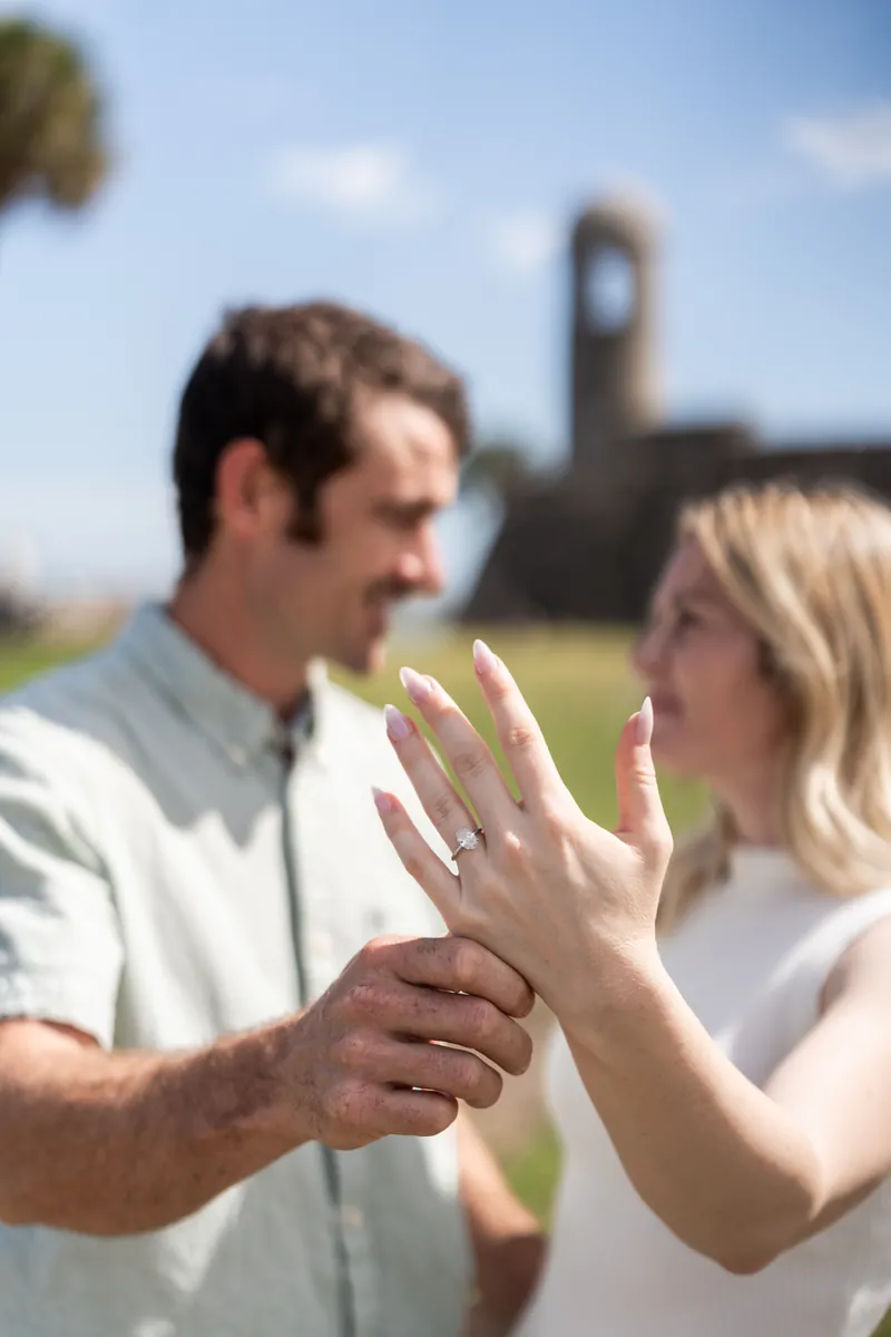 Dillon and Samantha hold hands and admire her engagement ring with the historic fort structure blurred in the background.