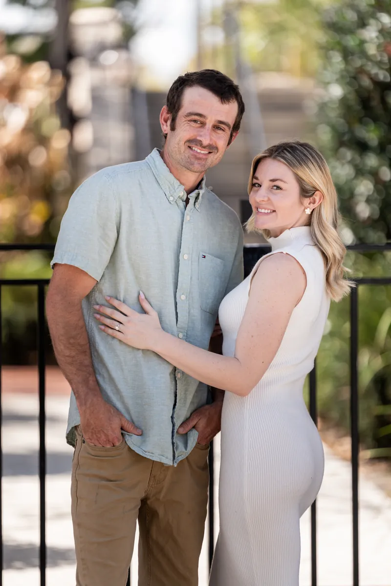 Samantha and Dillon stand close together on a wrought-iron balcony with ivy-covered railings and soft afternoon light.
