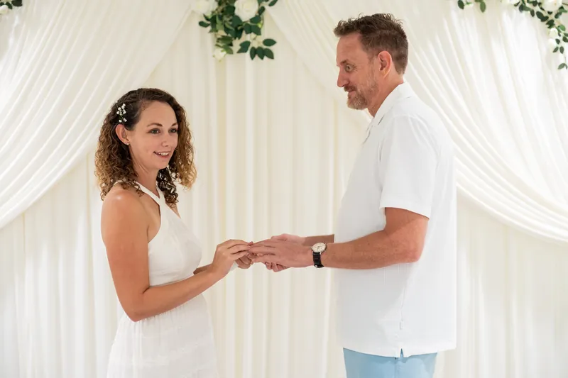 Daniel places a ring on Samantha's finger under white drapery with green garland.