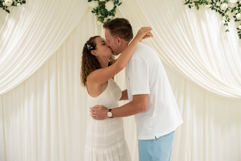 Samantha and Daniel kiss under the white curtain and floral arrangement, their arms wrapped around each other in celebration.