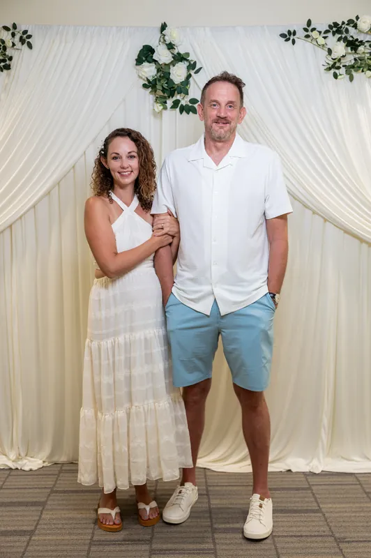 Samantha in a sleeveless white dress and Daniel in a white shirt and blue shorts stand together outdoors.