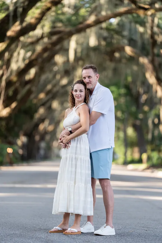 The couple stands arm-in-arm beneath sprawling oak branches with dappled light overhead.