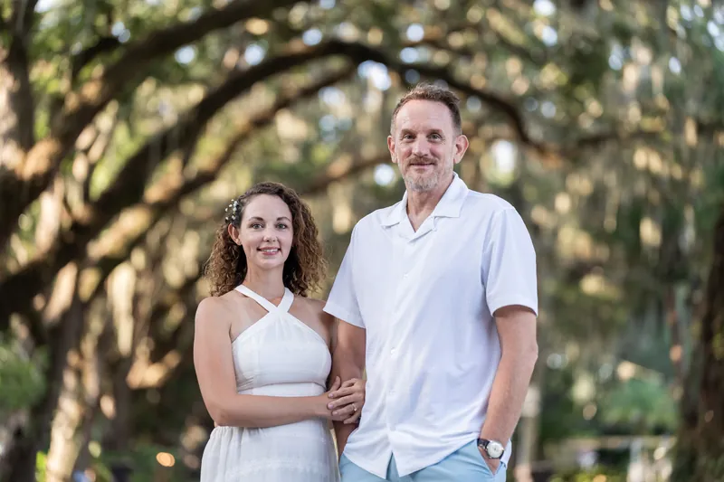 Samantha and Daniel embrace while walking under towering oaks in their white outfits.