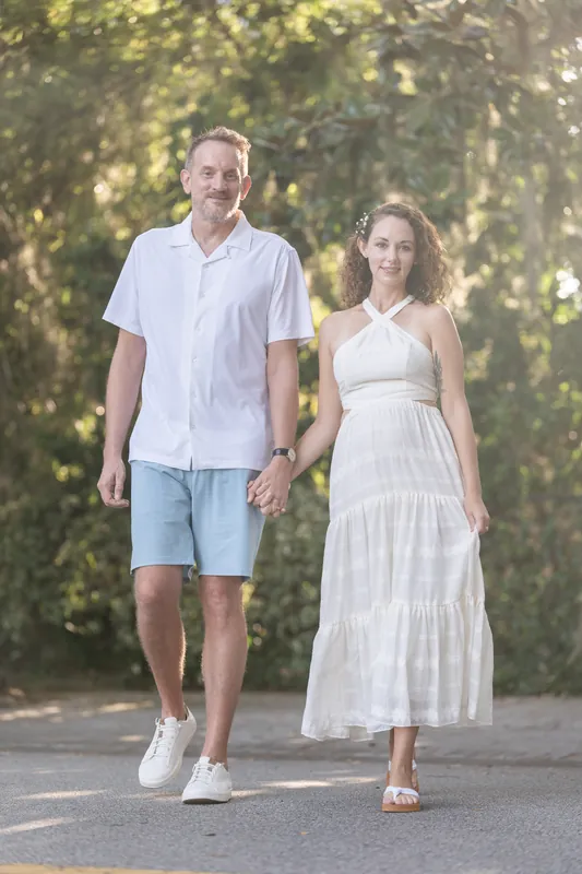 Walking hand in hand down a tree-lined path, the couple moves through dappled sunlight filtering through dense canopy.