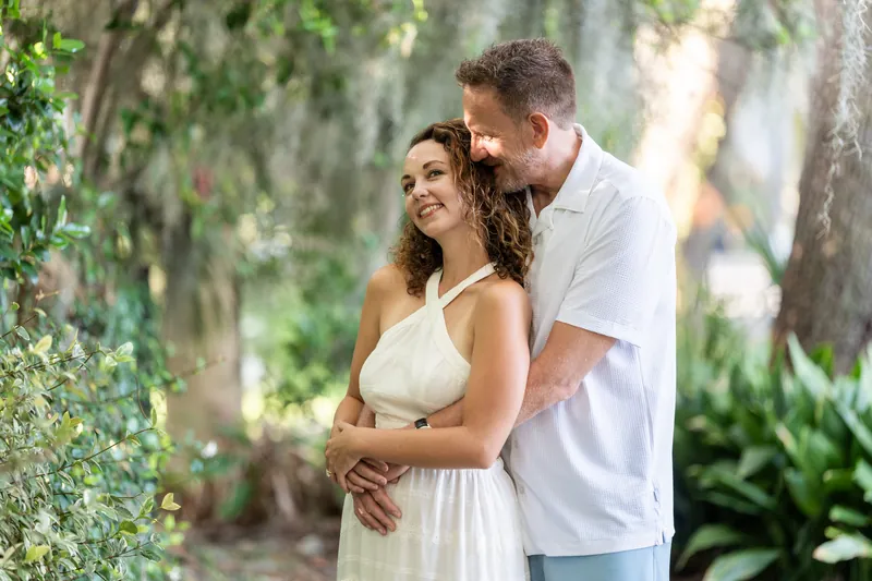 Daniel embraces Samantha from behind under the shade of hanging moss and oak branches, both smiling warmly.