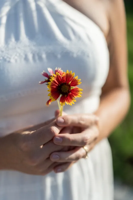 Samantha holds a vibrant red and yellow flower in her hands while wearing a white halter dress at Vilano Beach.