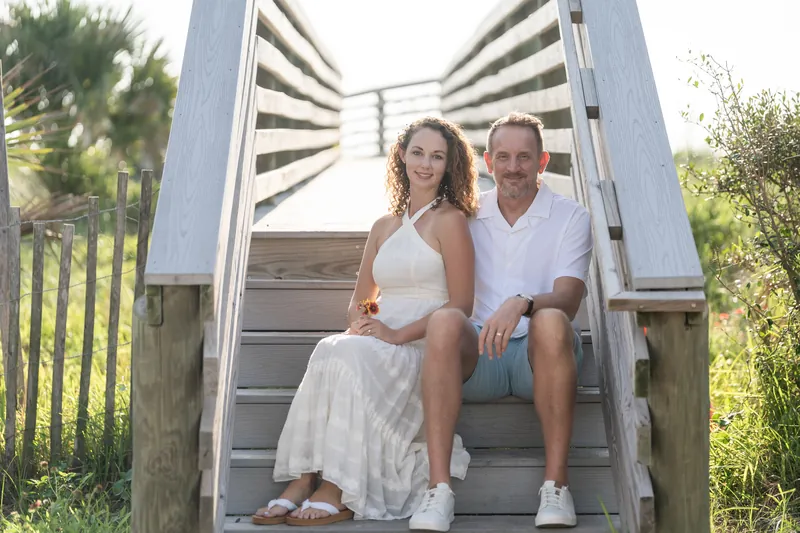 Samantha and Daniel sit together on wooden beach stairs beneath a striped umbrella, smiling in the afternoon sun.