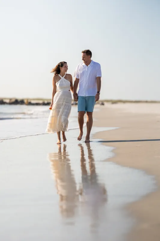 The couple walks barefoot through shallow water and wet sand along the shoreline, their reflections visible below them.