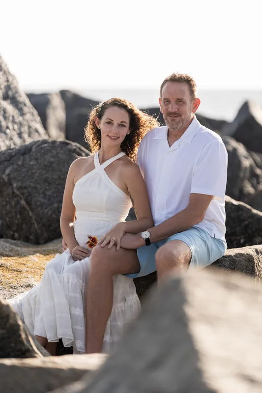 Daniel and Samantha sit close among large boulders on the beach, his arm around her shoulder as they gaze at each other.