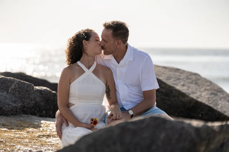 Leaning in for a kiss between dark rocks, the couple shares an intimate moment with the ocean visible behind them.