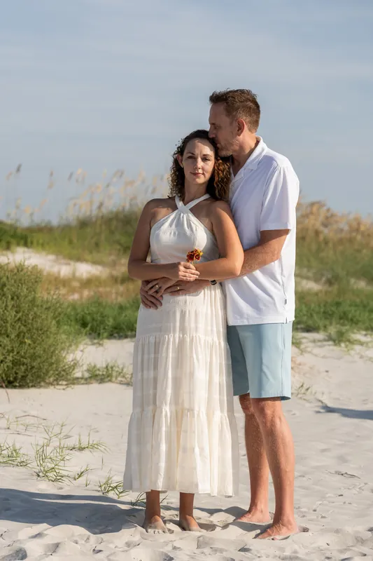 Samantha and Daniel stand together on sandy dunes with beach grass behind them at Vilano Beach, Daniel in a light blue shirt and shorts, Samantha in a cream linen dress.