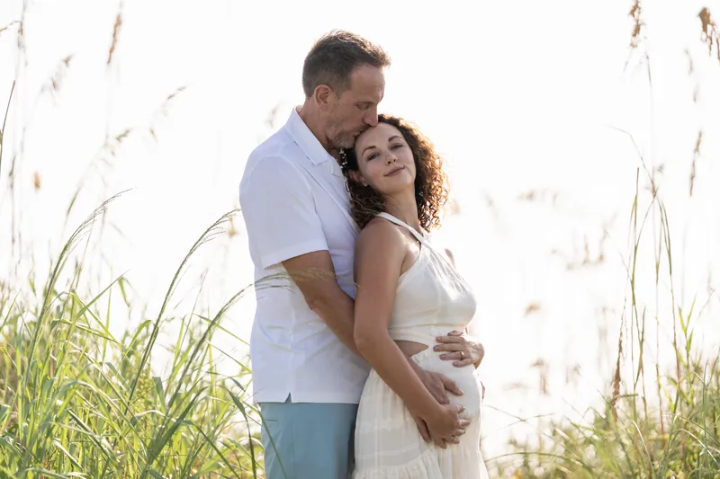 Daniel holds Samantha from behind near golden dune grasses, her curly hair catching light.