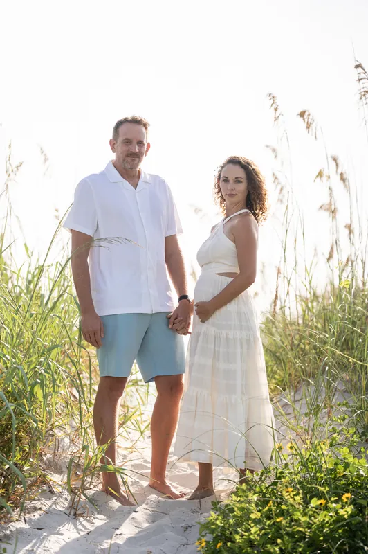 Samantha and Daniel walk hand-in-hand through coastal vegetation, her cream dress flowing beside his blue shorts.