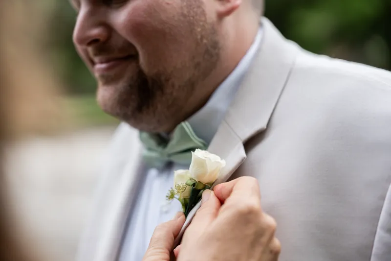 A hand pins a white and green boutonniere to Matthew's light blue shirt and gray jacket.