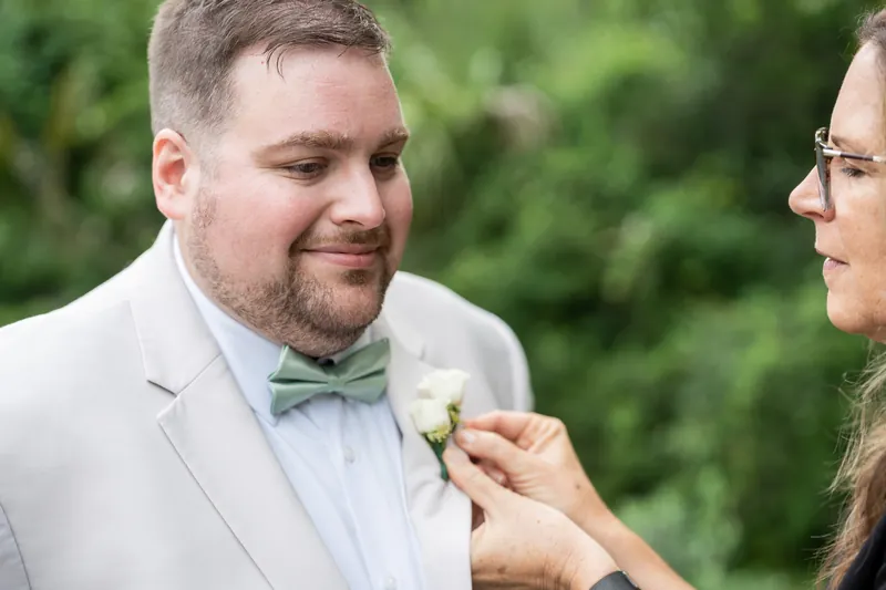 Samantha pins a white flower boutonniere onto Matthew's light blue shirt as he grins at the garden venue.