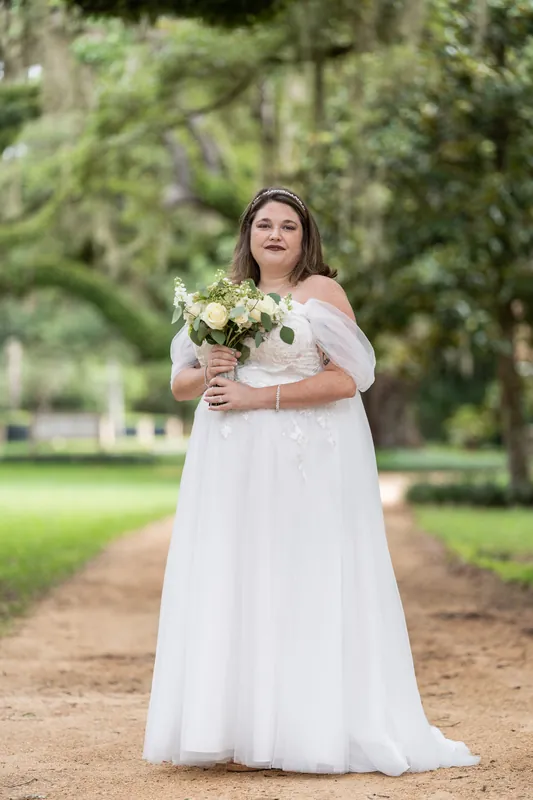 Samantha stands on a tree-lined path holding her bridal bouquet in a flowing white dress at Washington Oaks Gardens.