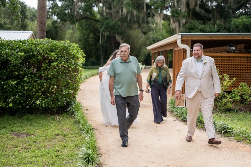 Matthew and two family members walk along a tree-lined path past manicured hedges and a wooden structure.