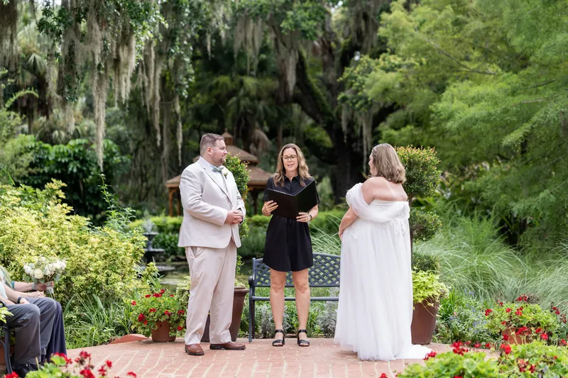 A woman in white and Matthew in cream listen to an officiant in black during their ceremony surrounded by flowering plants.