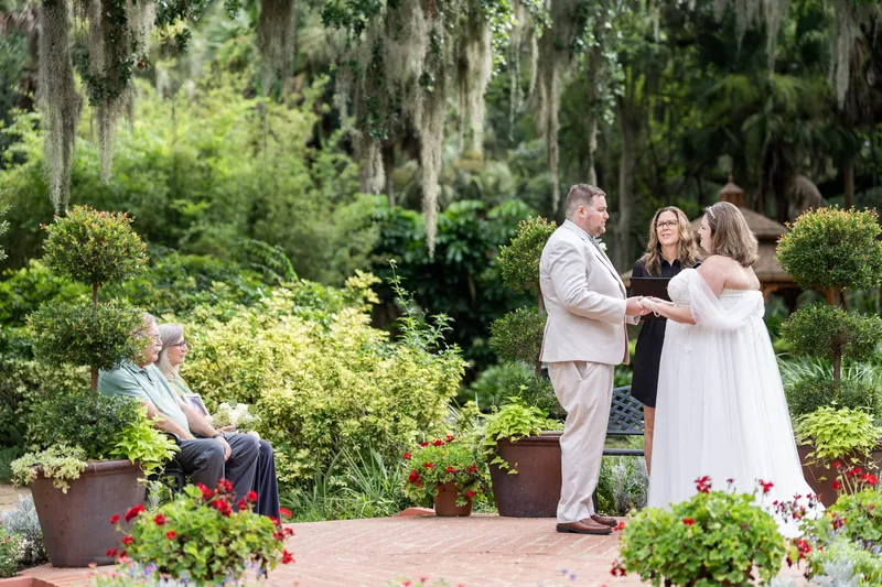 Matthew holds Samantha's hand during their ceremony as a guest sits nearby amid the lush, moss-laden landscape.