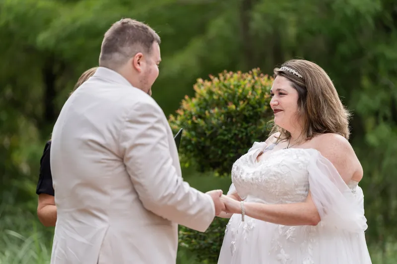Samantha gazes at Matthew while holding his hand in her flowing white dress against a soft green garden backdrop.