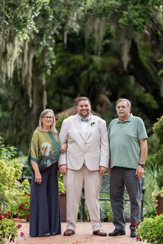 Matthew stands between two family members on the brick path, surrounded by lush green foliage and flowering plants.