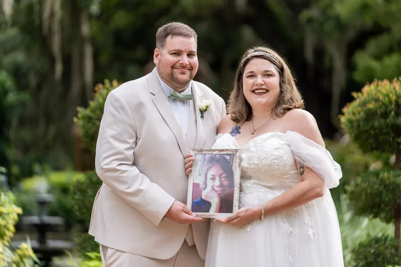 Samantha and Matthew hold a framed photo together, both smiling beneath the canopy of ancient oaks and lush greenery.