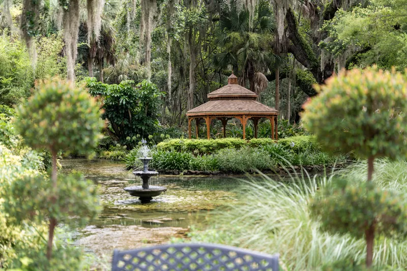 A wooden gazebo reflected in still water, surrounded by manicured gardens and moss-laden trees in soft afternoon light.