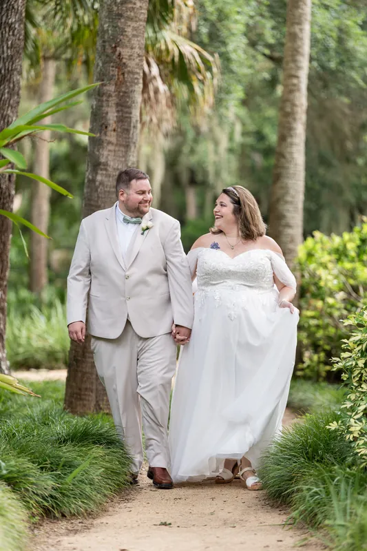 Samantha in a white off-shoulder gown and Matthew in cream walk hand-in-hand down a shaded garden path.