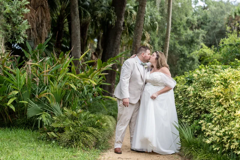 Matthew kisses Samantha's forehead as they embrace beneath towering oaks and lush garden growth.