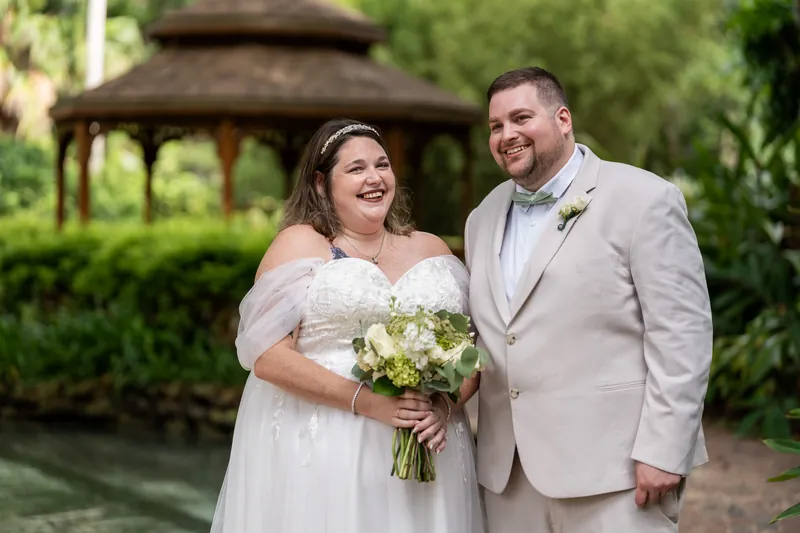Matthew and Samantha smile together as she holds a white bridal bouquet, a wooden gazebo visible behind them.