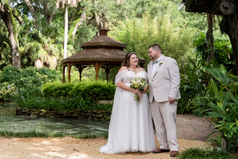 The couple stands hand-in-hand near a pond with a gazebo, surrounded by manicured gardens and trees.