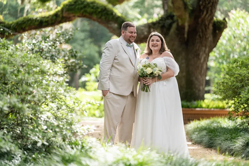Samantha and Matthew stand together under sprawling oak trees at Washington Oaks Gardens, with Samantha holding a white bouquet in her cream dress.