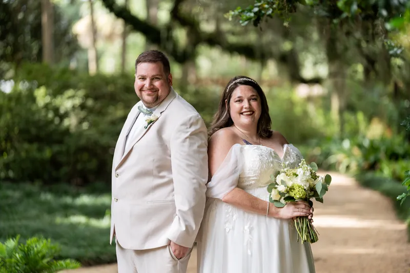 Matthew and Samantha walk side by side down a shaded garden path, both smiling at the camera as Spanish moss drapes overhead.